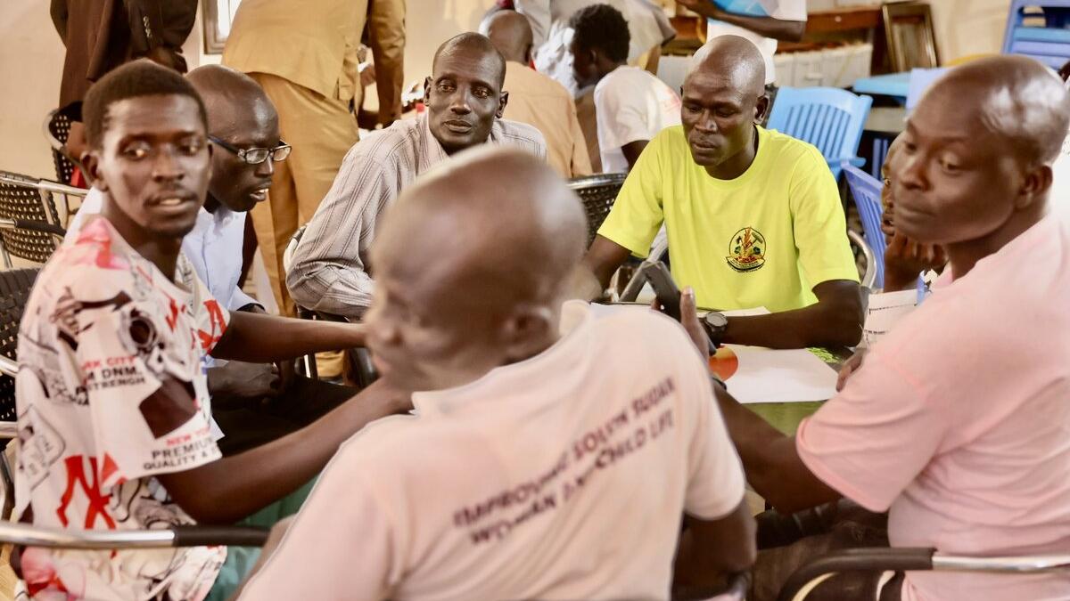 Several people seated around tables indoors, engaged in discussion, with chairs and documents visible in the background.
