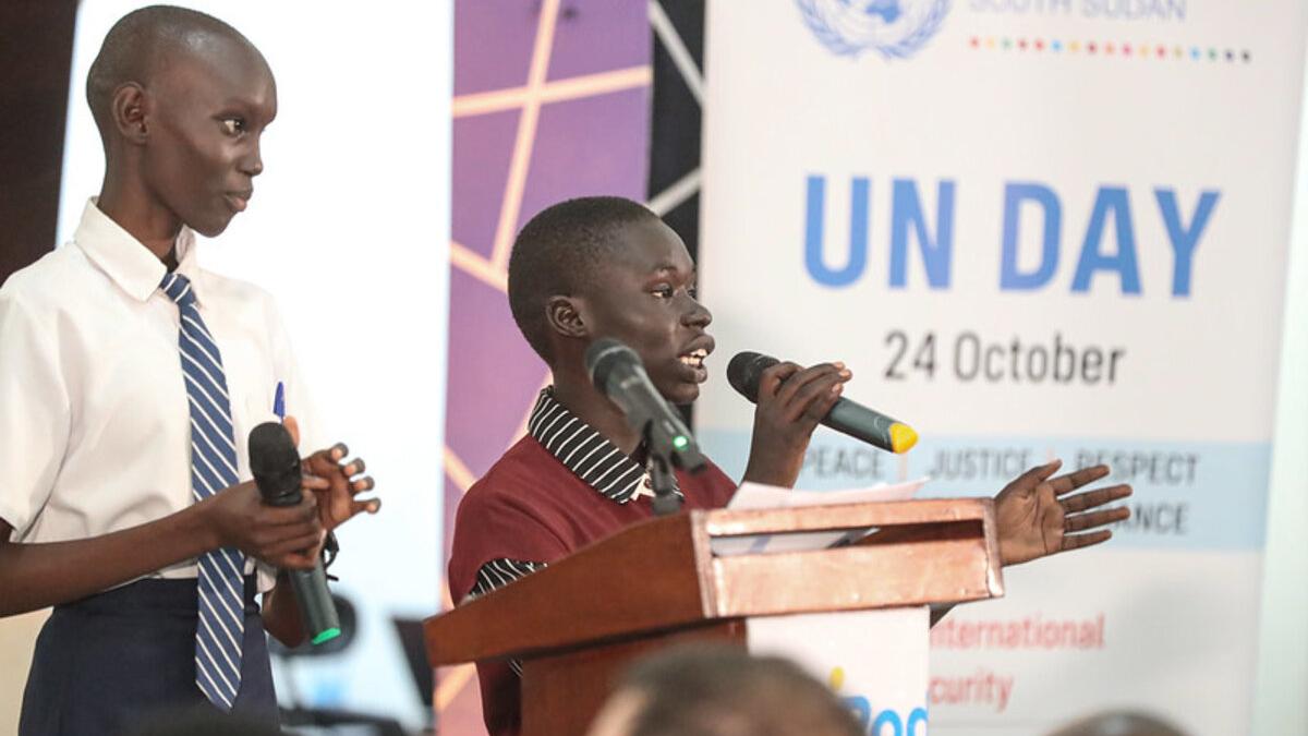 Two individuals speaking at a podium with microphones during a UN Day event, with a banner showing ‘UN Day 24 October’ in the background.
