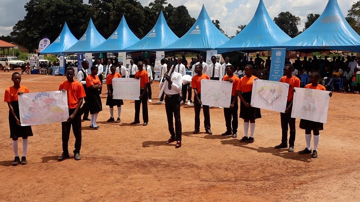 Students in orange shirts and black skirts or trousers hold large hand-drawn posters during an outdoor event with blue tents and a crowd in the background.