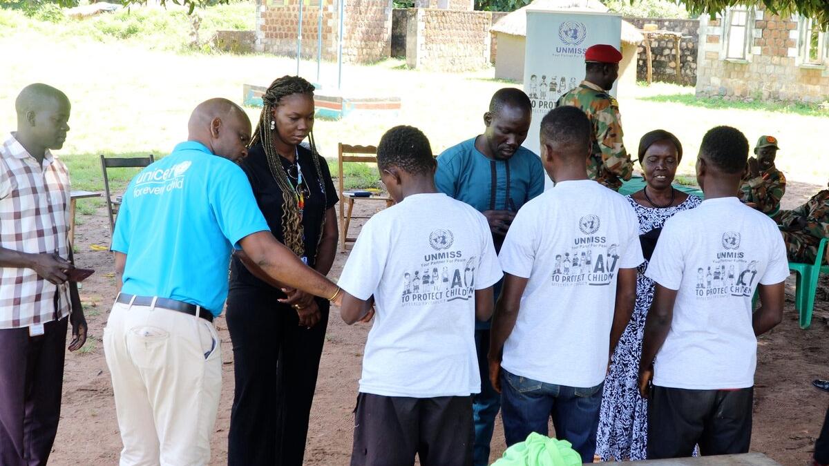 Group of people standing outdoors in a circle, including individuals wearing UNMISS-branded shirts and a person in a UNICEF shirt, with soldiers and chairs in the background.