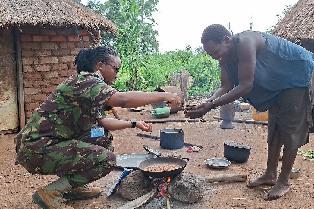 A person in camouflage uniform kneels beside an outdoor cooking area, handing something to another person near a fire with pots and a pan in front of traditional huts